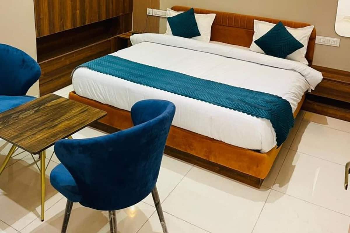Una habitación de hotel moderna con un armario de madera, una cama con ropa de cama blanca y aguamarina, dos sillas azules e iluminación colgante.