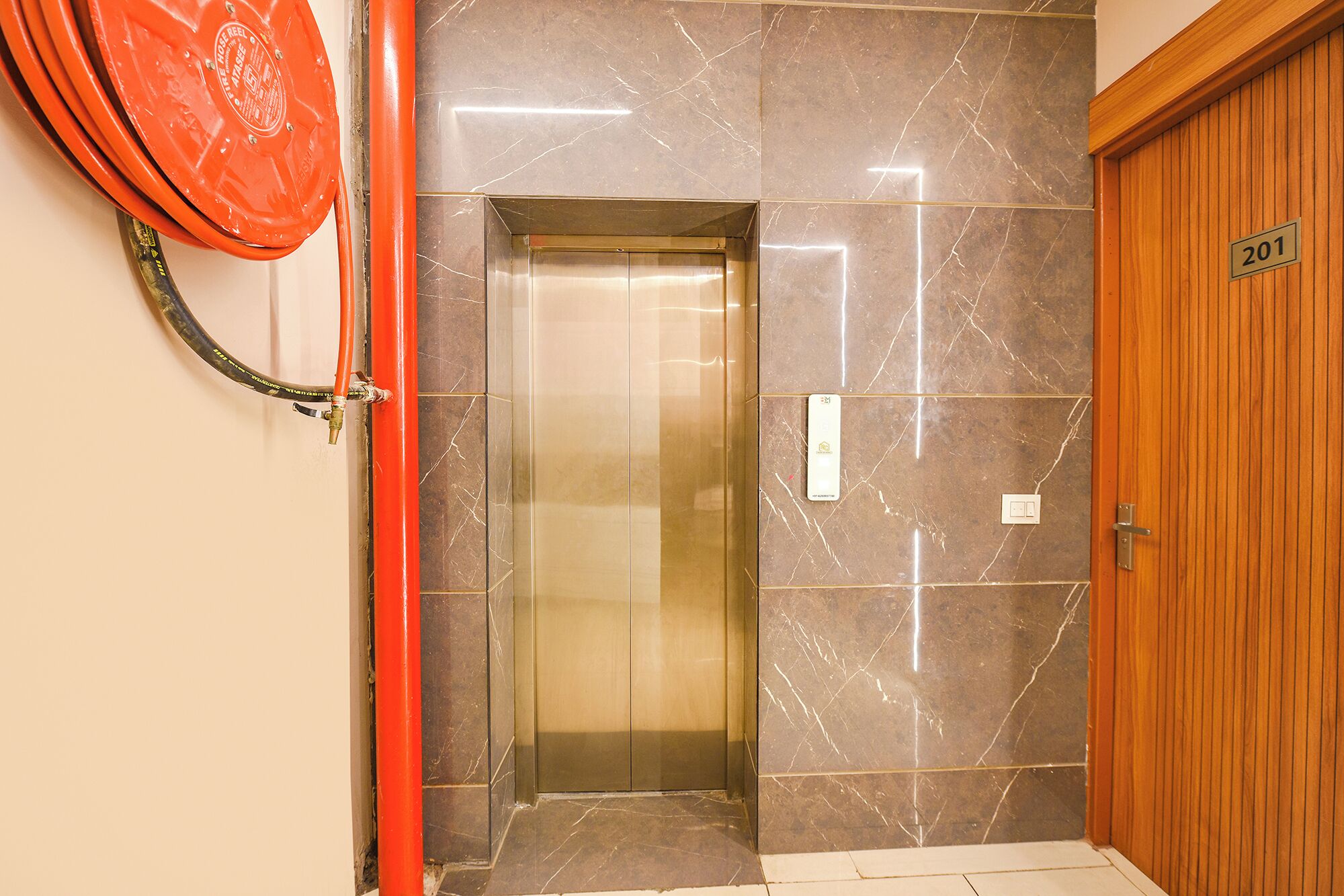 Un ascenseur d’hôtel avec des murs en marbre, une porte en bois portant une plaque avec un numéro, et un tuyau rouge.
