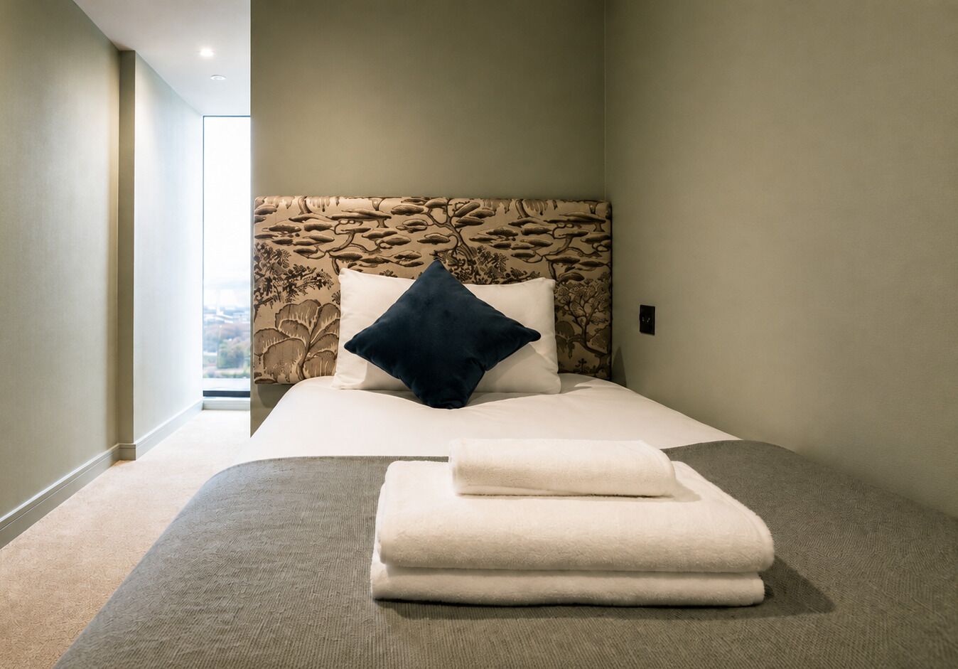 A hotel room with a bed, a headboard with a leaf pattern, a window, and a view of the outside.
