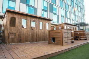 A wooden cabin-style building with large windows, a wooden deck, and a modern building in the background. - Southall &Soul (Hayes)