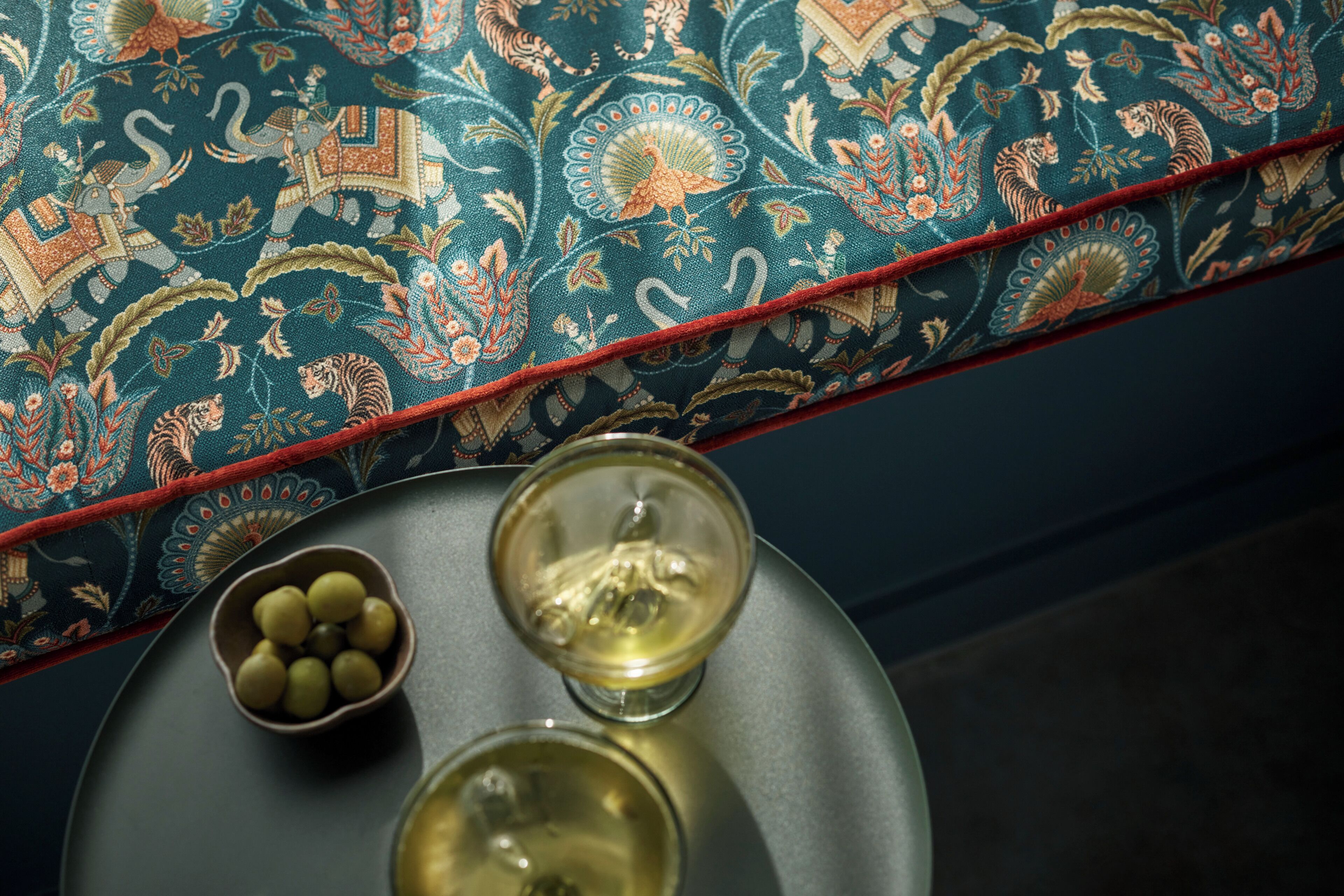 A round table with a bowl of olives and two glasses of white wine, set against a patterned fabric backdrop.