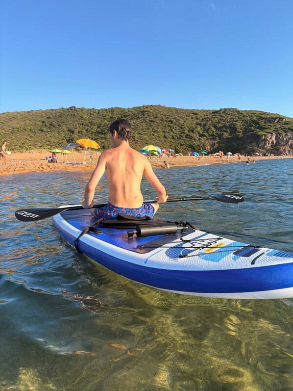 Una persona rema en un kayak azul sobre aguas claras, con una playa y sombrillas al fondo.