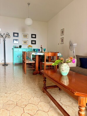 Un coin repas avec une table en bois, des chaises, un vase avec des fleurs, une cuisine en arrière-plan et un ventilateur sur la droite.