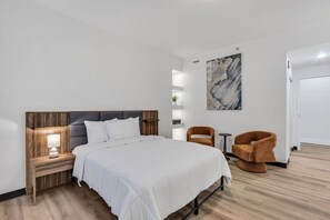 A modern hotel room with a large bed, wooden headboard, bedside tables with lamps, a small side table, a brown armchair, a round table, and a wall-mounted shelf with a plant.