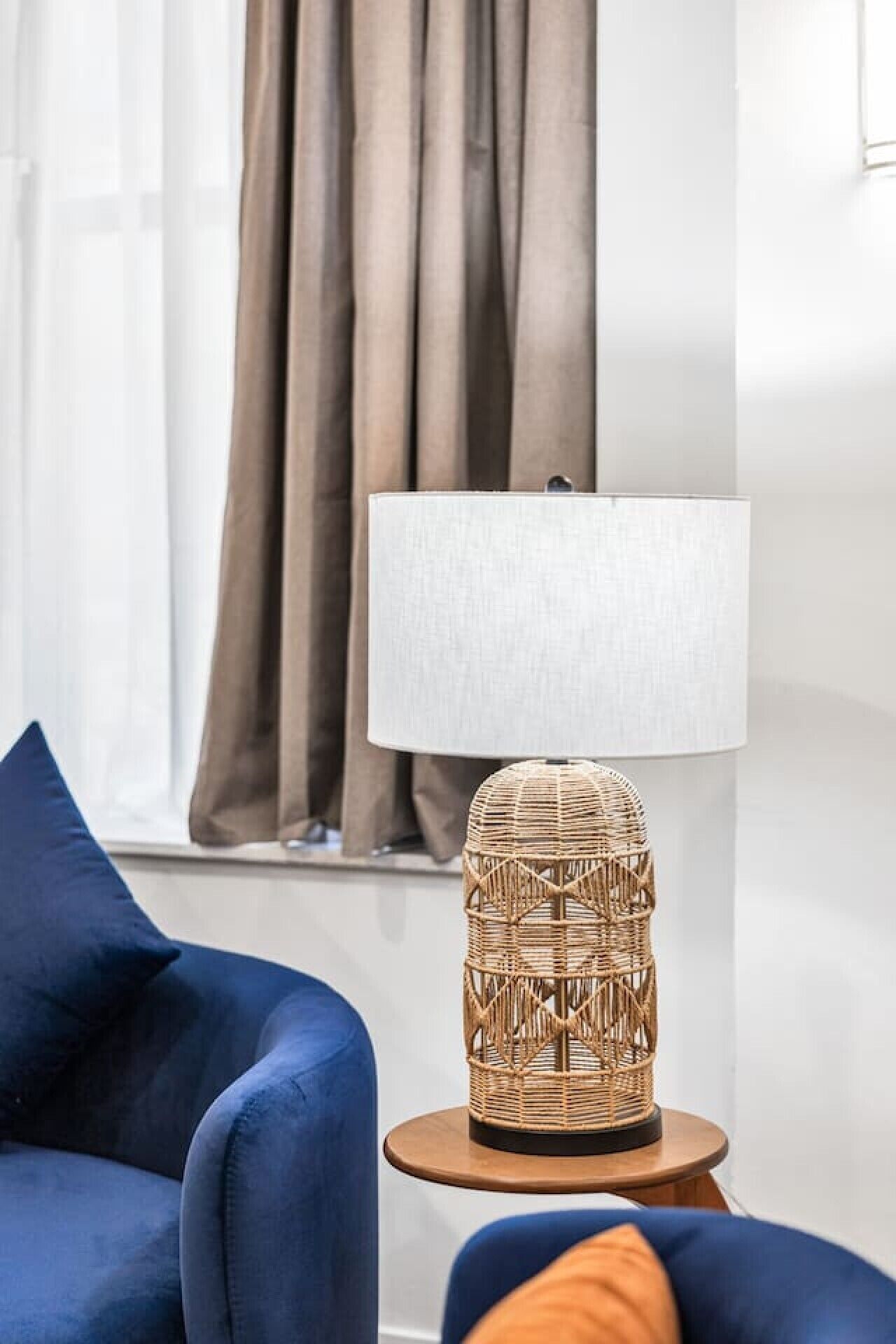 Ein Zimmer mit einem blauen Sofa, einer Korblampe auf einem Holztischchen und einem Fenster mit Vorhängen.