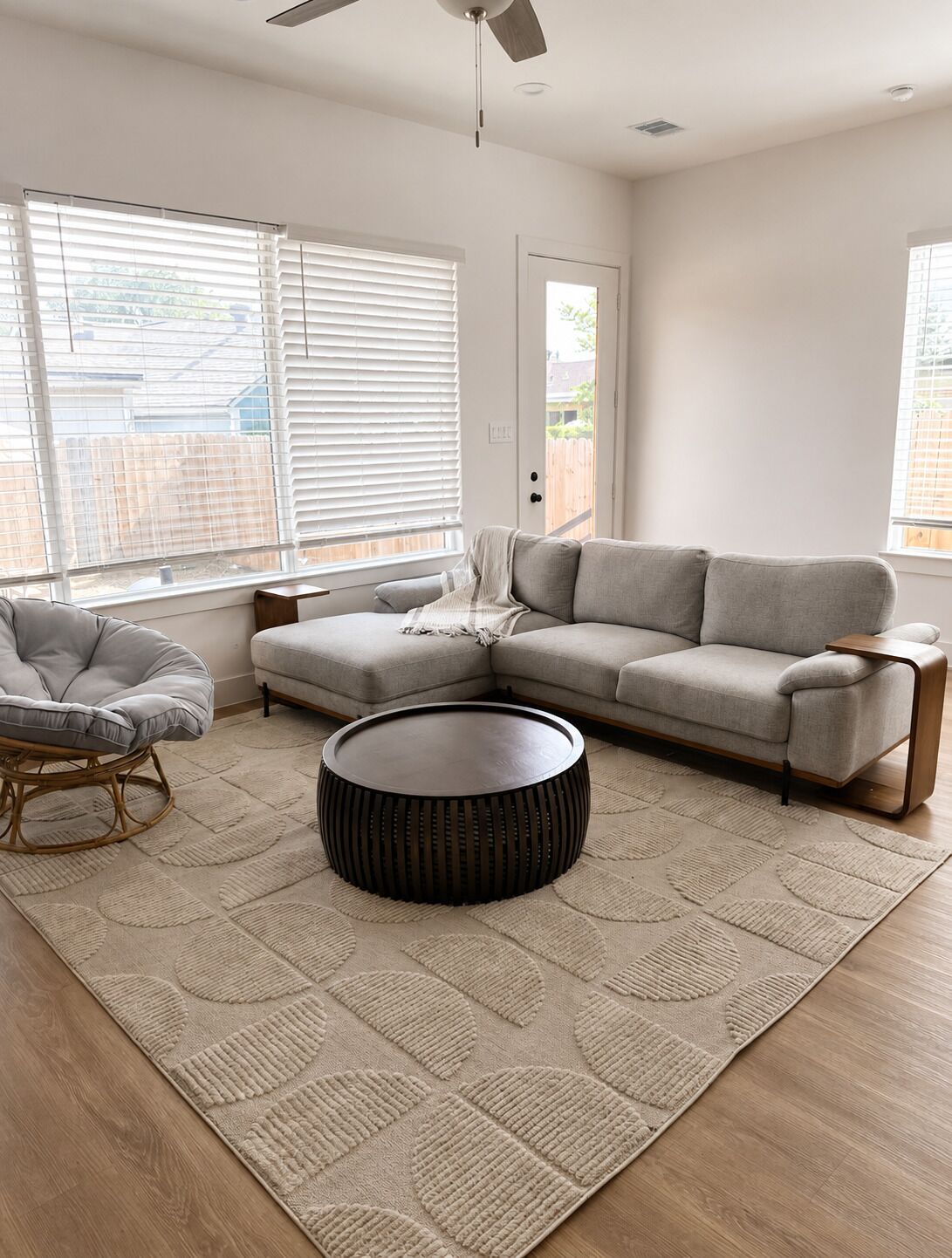 Un salon comprenant un canapé, une table basse ronde, un fauteuil et une grande fenêtre avec des stores.