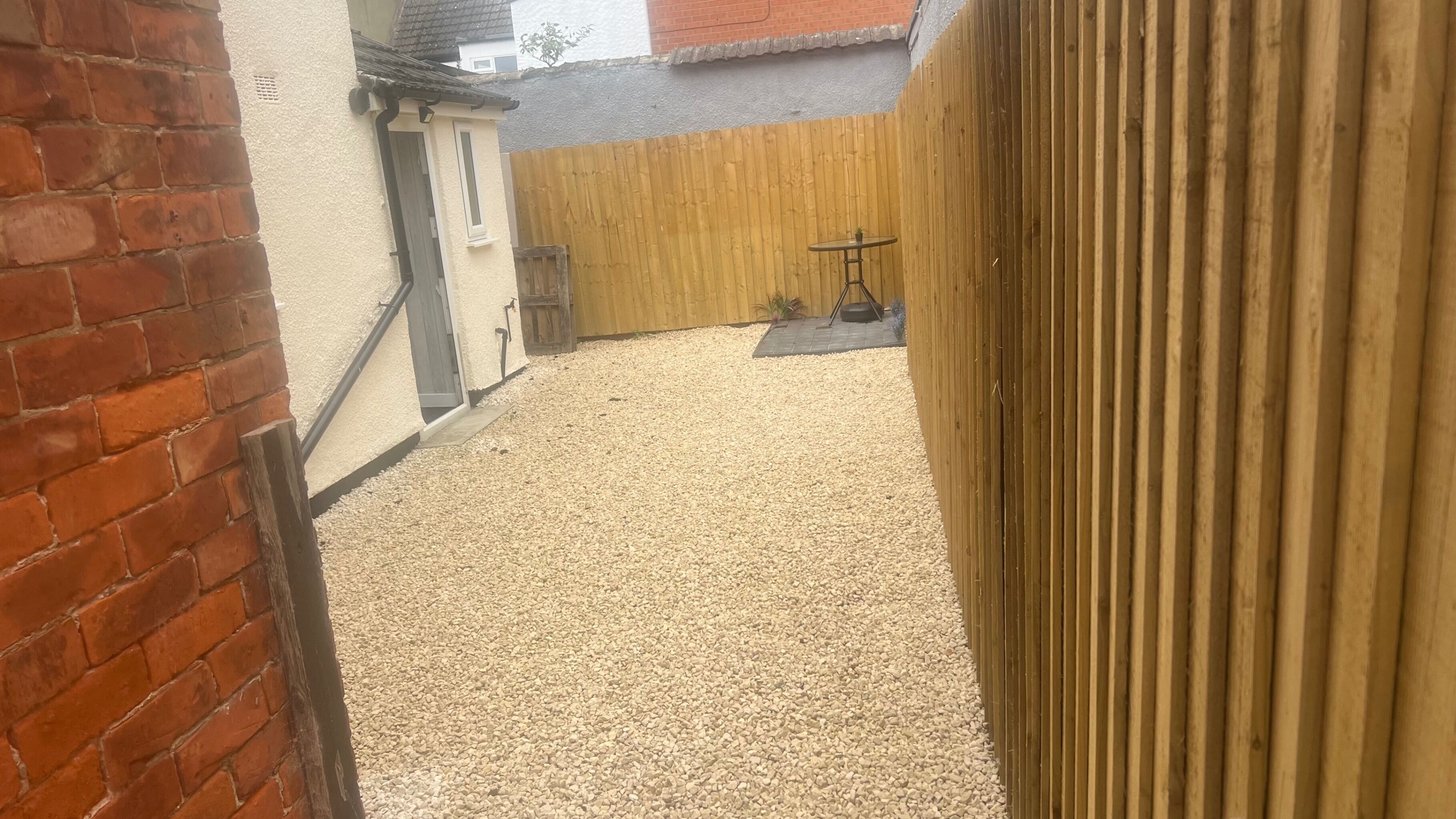 Ein Garten mit Kies, einem Holenzaun und einem kleinen Tisch.