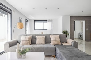 Ein modernes Wohnzimmer mit einem grauen Ecksofa, einem weißen Couchtisch und einem großen Fenster mit Vorhängen.