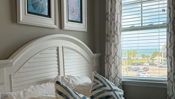 Une chambre à coucher avec un lit, deux tableaux encadrés, une fenêtre avec des rideaux et une vue sur l’extérieur.
