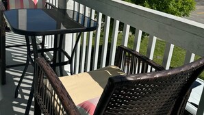 Un balcon avec des fauteuils en osier et une table, donnant sur une rue bordée d’immeubles et d’arbres.