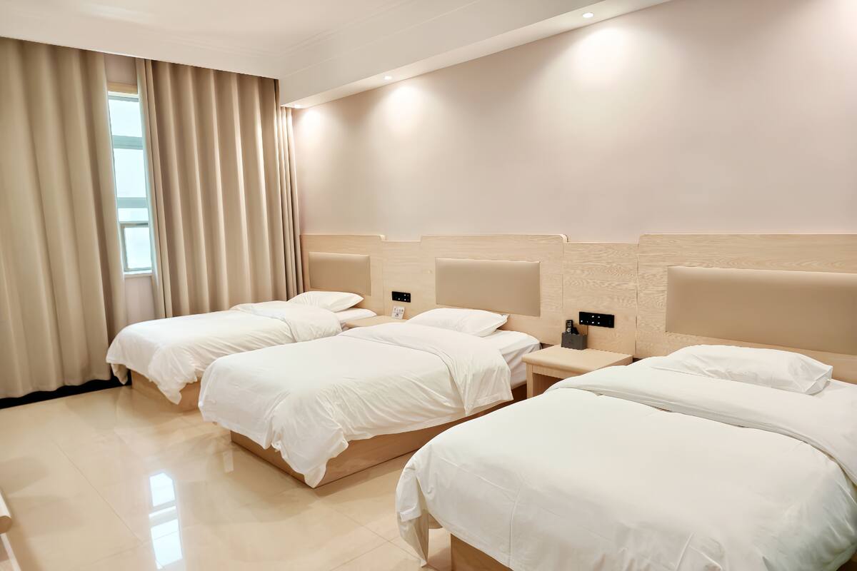 Ein Hotelzimmer mit drei Betten, einem Holz-Kopfteil, einem Nachttisch mit Lampe und einem Fenster mit Vorhängen.