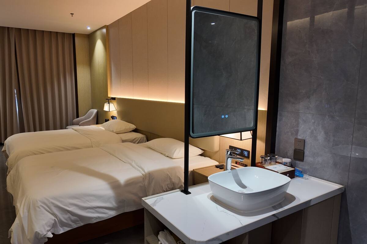 Ein Hotelzimmer mit zwei Betten, einem Nachttisch mit Waschbecken und einer fest installierten Bedienanlage sowie einem großen Spiegel.