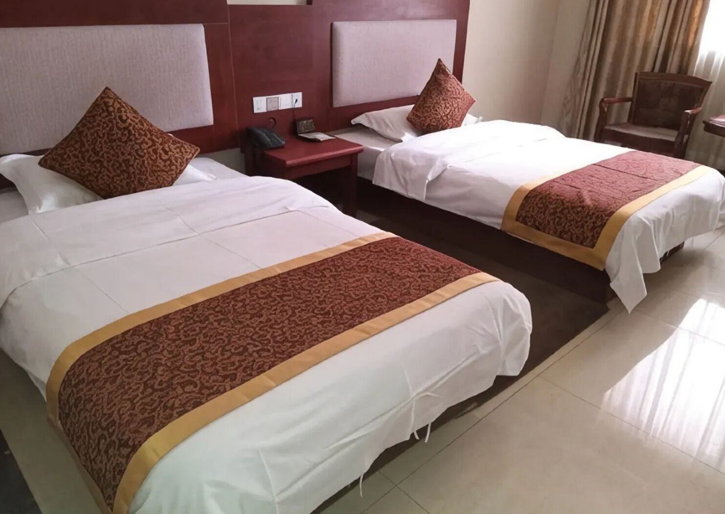 A hotel room with two beds, a nightstand, a chair, and a window with curtains.