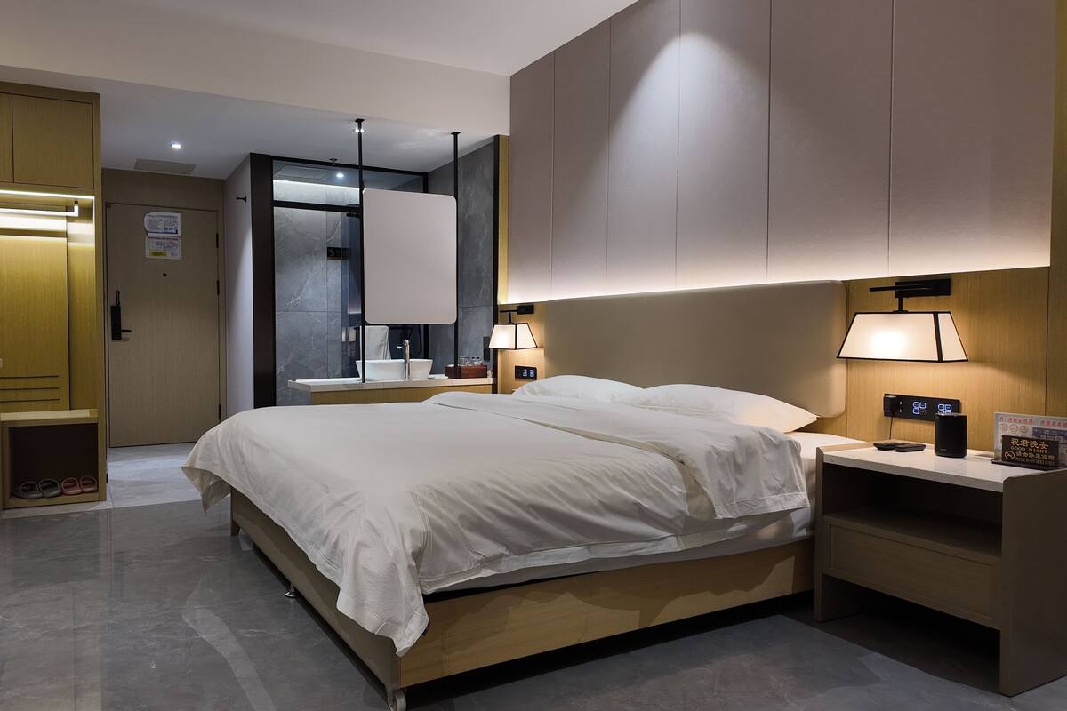 Ein modernes Hotelzimmer mit einem großen Bett, Nachttischen, einem Spiegel, einem Kleiderschrank und einem durchgegangene Tür sichtbaren Badezimmer.
