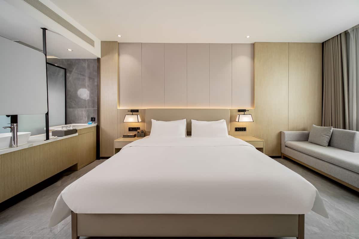 Ein modernes Hotelzimmer mit einem großen Bett, Nachttischlampen, einem Sofa, einem Schminktisch mit Spiegel und einem großen Fenster mit Vorhängen.