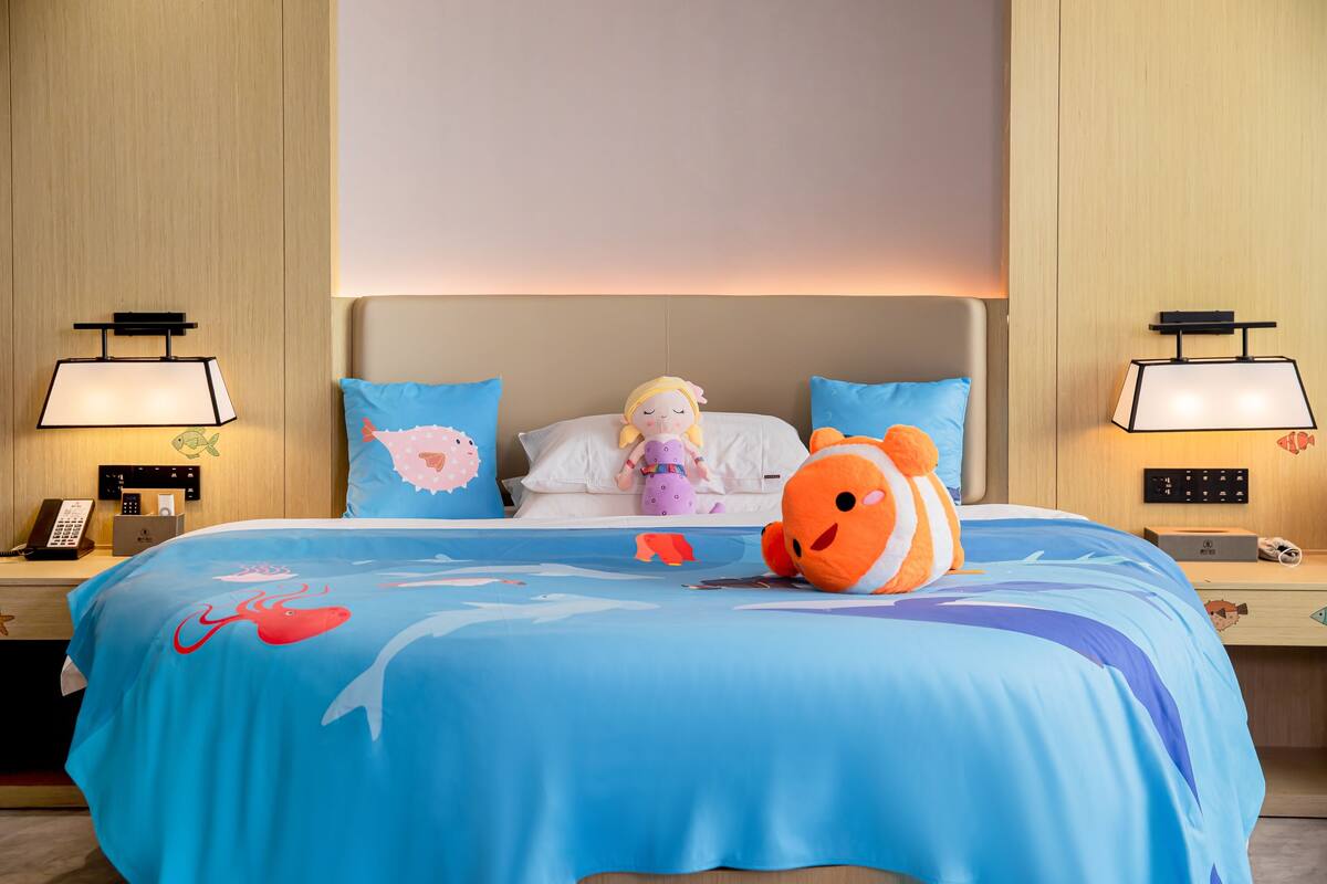 Ein Hotelzimmer mit einem Bett, zwei Nachttischen, einer Lampe, einem Kuscheltier und einem Kissen mit Fischmuster.