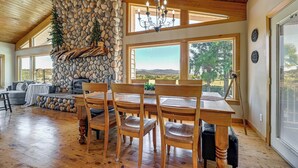 A spacious room with a stone fireplace, wooden ceiling, and large windows offering a view of the outdoors. - Private Mtn-view Pool! Rogue River Valley Escape (Eagle Point)