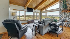A spacious living room with a high wooden ceiling, large windows offering a view of the countryside, a stone fireplace, and leather seating. - Private Mtn-view Pool! Rogue River Valley Escape (Eagle Point)