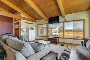 A cozy living room with a wooden ceiling, large windows, a sofa, a table, and a view of a white car outside. - Private Mtn-view Pool! Rogue River Valley Escape (Eagle Point)