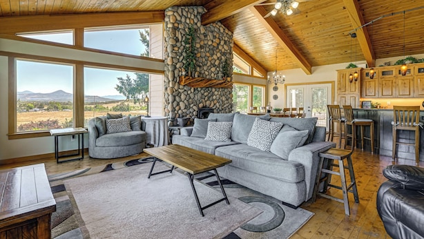 A spacious living area with a stone fireplace, large windows, and a kitchen with wooden cabinets. - Private Mtn-view Pool! Rogue River Valley Escape (Eagle Point)