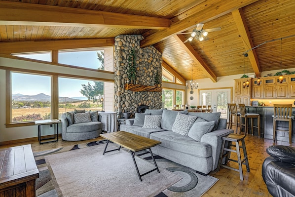 A spacious living area with a stone fireplace, large windows, and a kitchen with wooden cabinets. - Private Mtn-view Pool! Rogue River Valley Escape (Eagle Point)