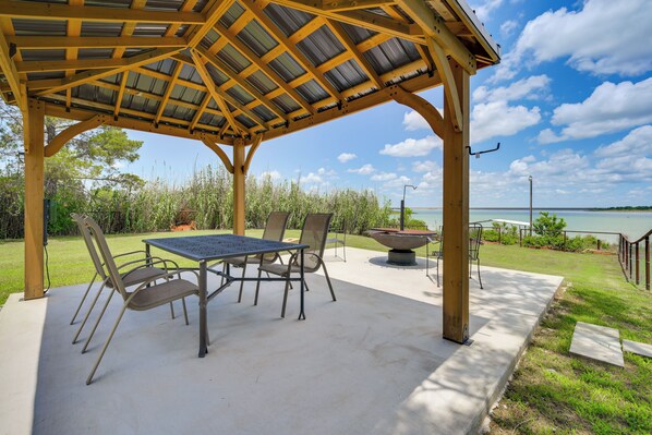 A covered outdoor patio with a table and chairs, a fire pit, and a view of a body of water.
