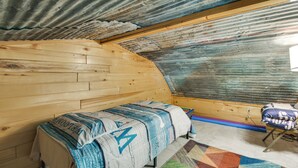 Una habitación con un banco de madera, una manta estampada, un tapete colorido y un techo de metal corrugado.