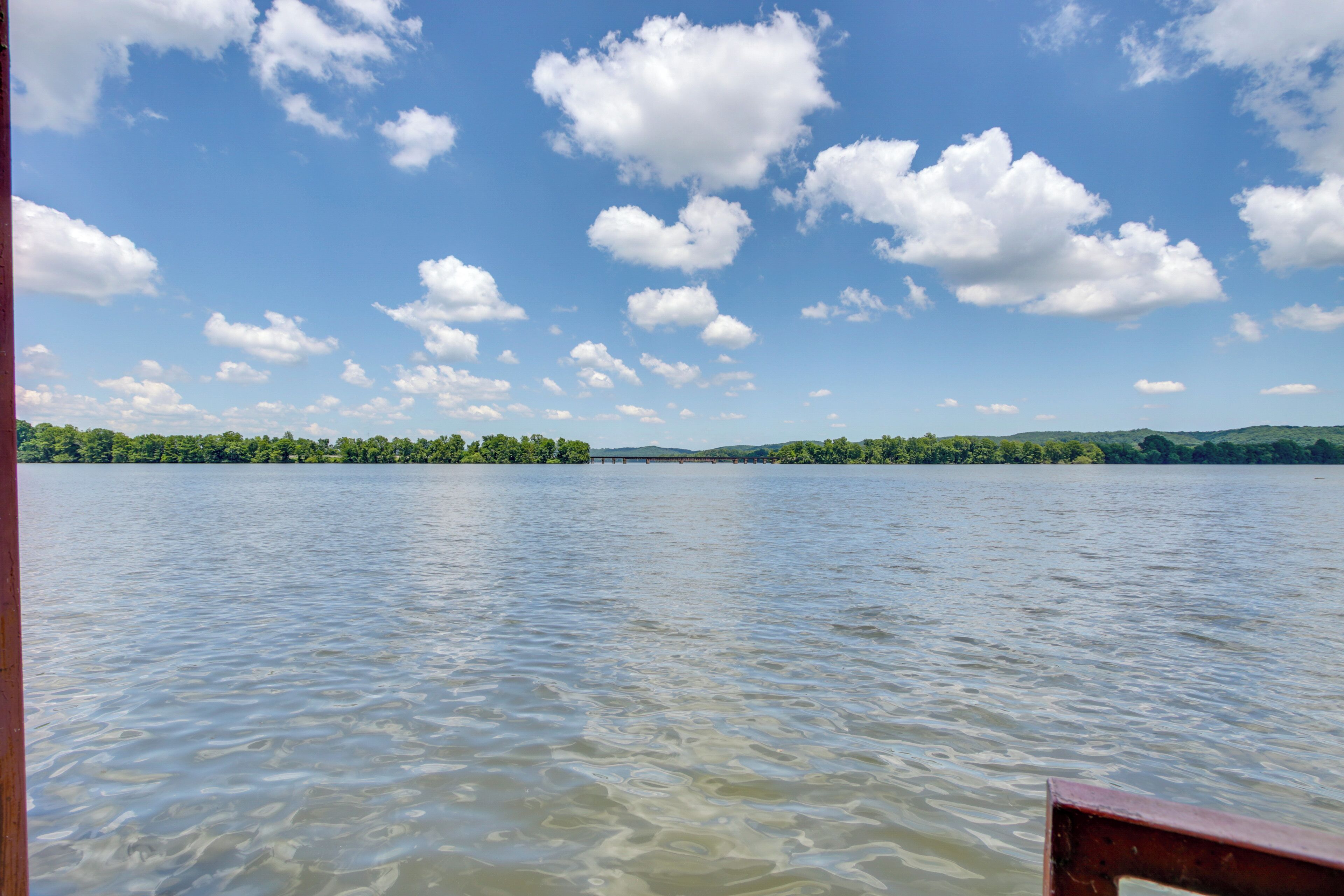 A calm lake with a wooden dock, surrounded by green hills and a clear blue sky with clouds.