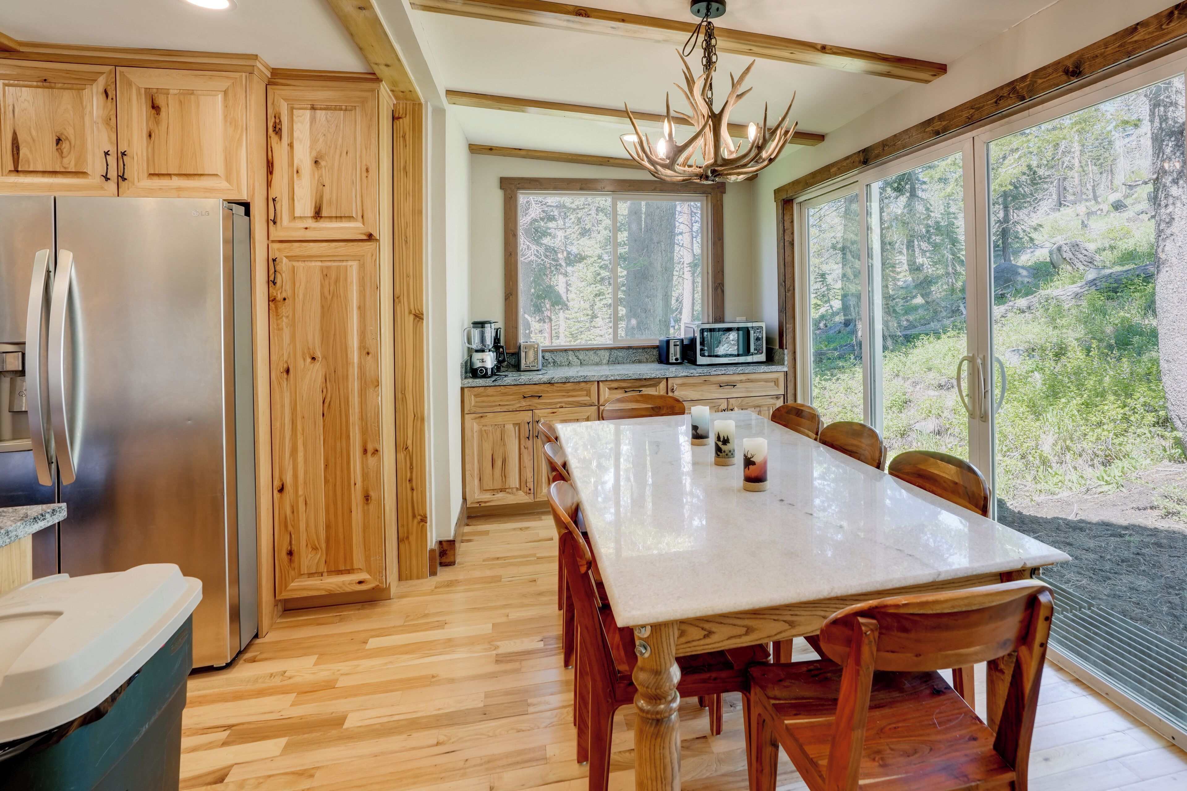 A modern kitchen with wooden cabinets, a stainless steel refrigerator, a dining table with chairs, and a large window with a forest view.