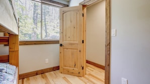 A room with a wooden door, a window with a forest view, a bed with a patterned bedspread, and a wooden floor. - Hike, Ski & Explore! Spacious Home in Twin Bridges (Echo Lake)