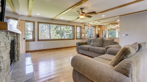 A spacious living room with a stone fireplace, wooden beams, large windows, and a ceiling fan. - Hike, Ski & Explore! Spacious Home in Twin Bridges (Echo Lake)