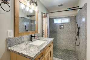 A bathroom with a granite countertop sink, wooden cabinets, a mirror, a shower with a glass door, and a window.