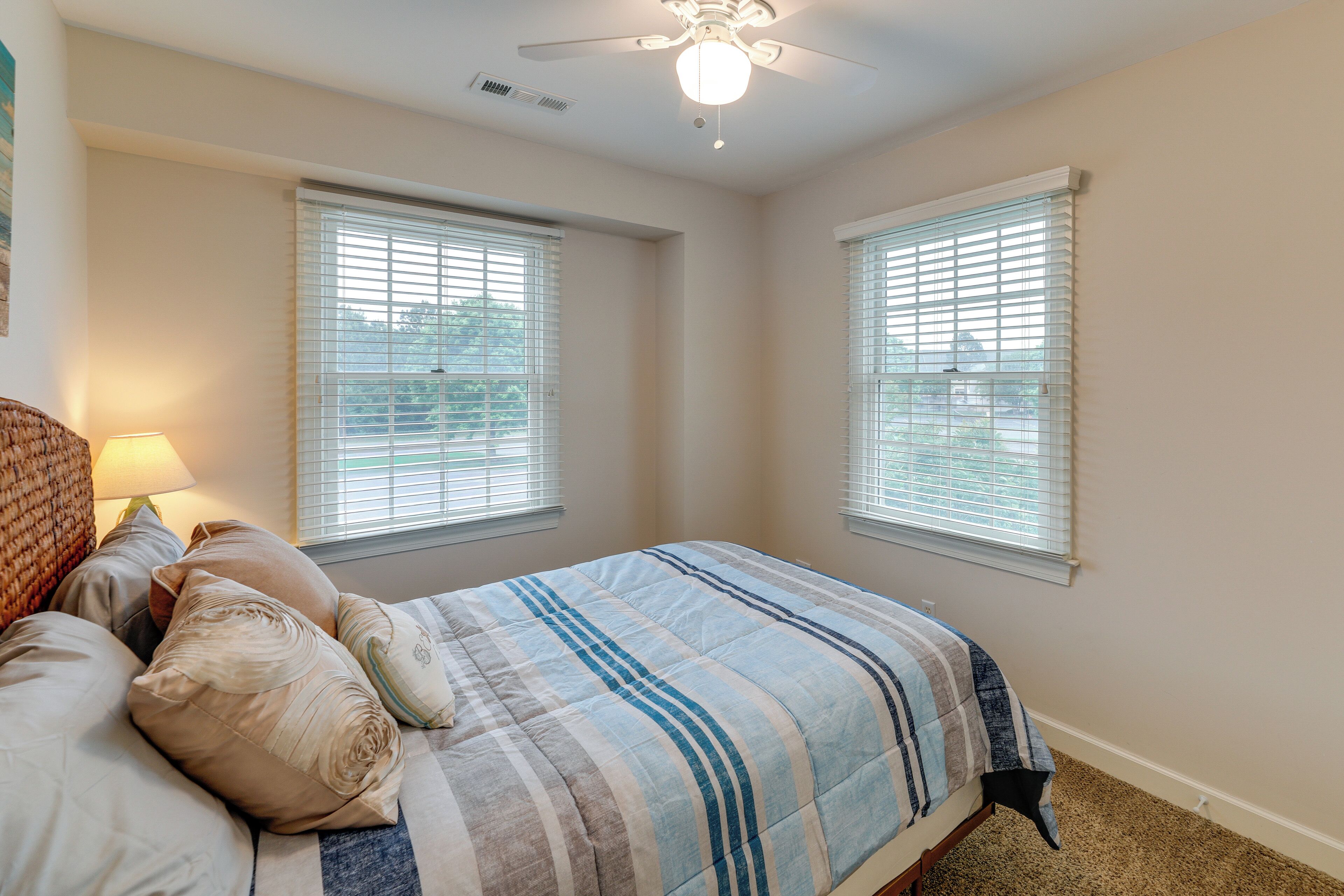 A bedroom with a bed, two windows with blinds, a ceiling fan, and a carpeted floor.