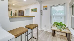 Une cuisine avec un plan de travail, deux tabourets, une fenêtre avec des stores et une plante en pot.