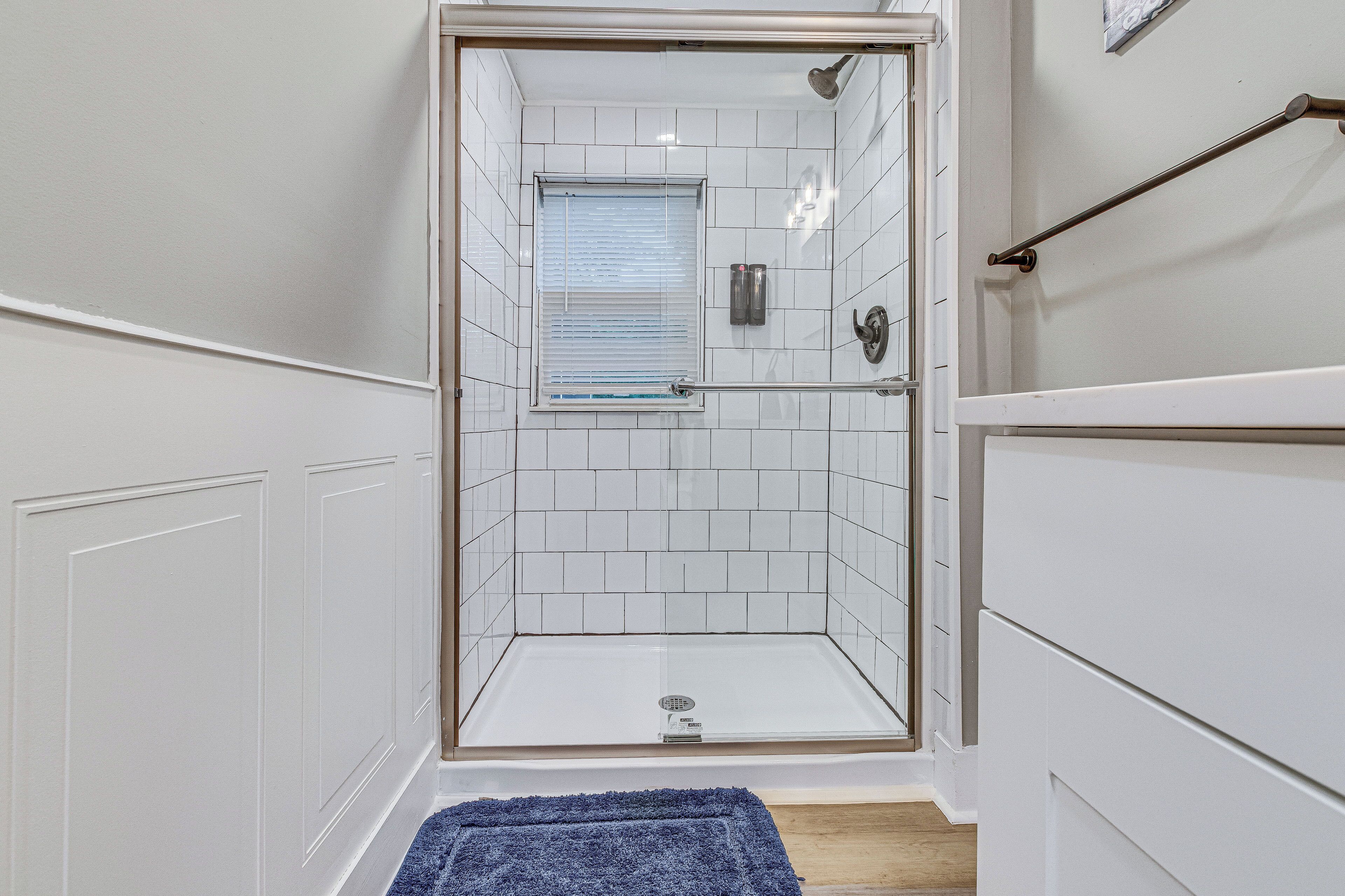 Um chuveiro com paredes de vidro, um revestimento de azulejos brancos, uma janela com persianas e um tapete de banho azul no chão.