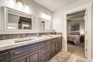 A bathroom with a double sink vanity, large mirrors, and a doorway leading to a bedroom.