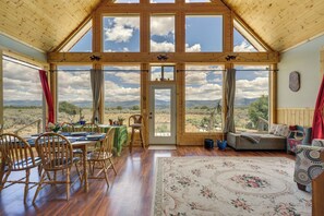 Ein geräumiger Raum mit hohen Decken, großen Fenstern, einer Essecke mit Holzsesseln, einem Sofa und einem Teppich mit Blumenmuster.