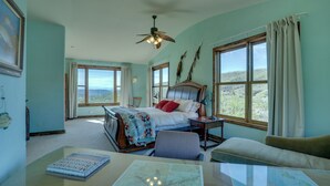 A bedroom with a bed, a desk with a map, a chair, a ceiling fan, and large windows.
