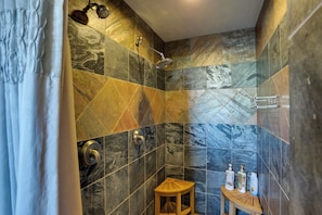 Une douche Ă©quipĂ©e dâun pommeau de douche Ă main, un tabouret en bois et des articles de toilette disposĂ©s sur une petite table.