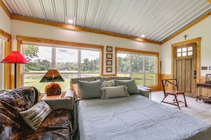 A bedroom with a large bed, a wooden ceiling, a window with a view of trees, and a door with a cross.