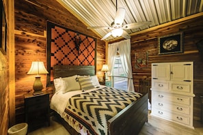 A wooden cabin bedroom with a bed, nightstands, a dresser, a ceiling fan, and a window with curtains.