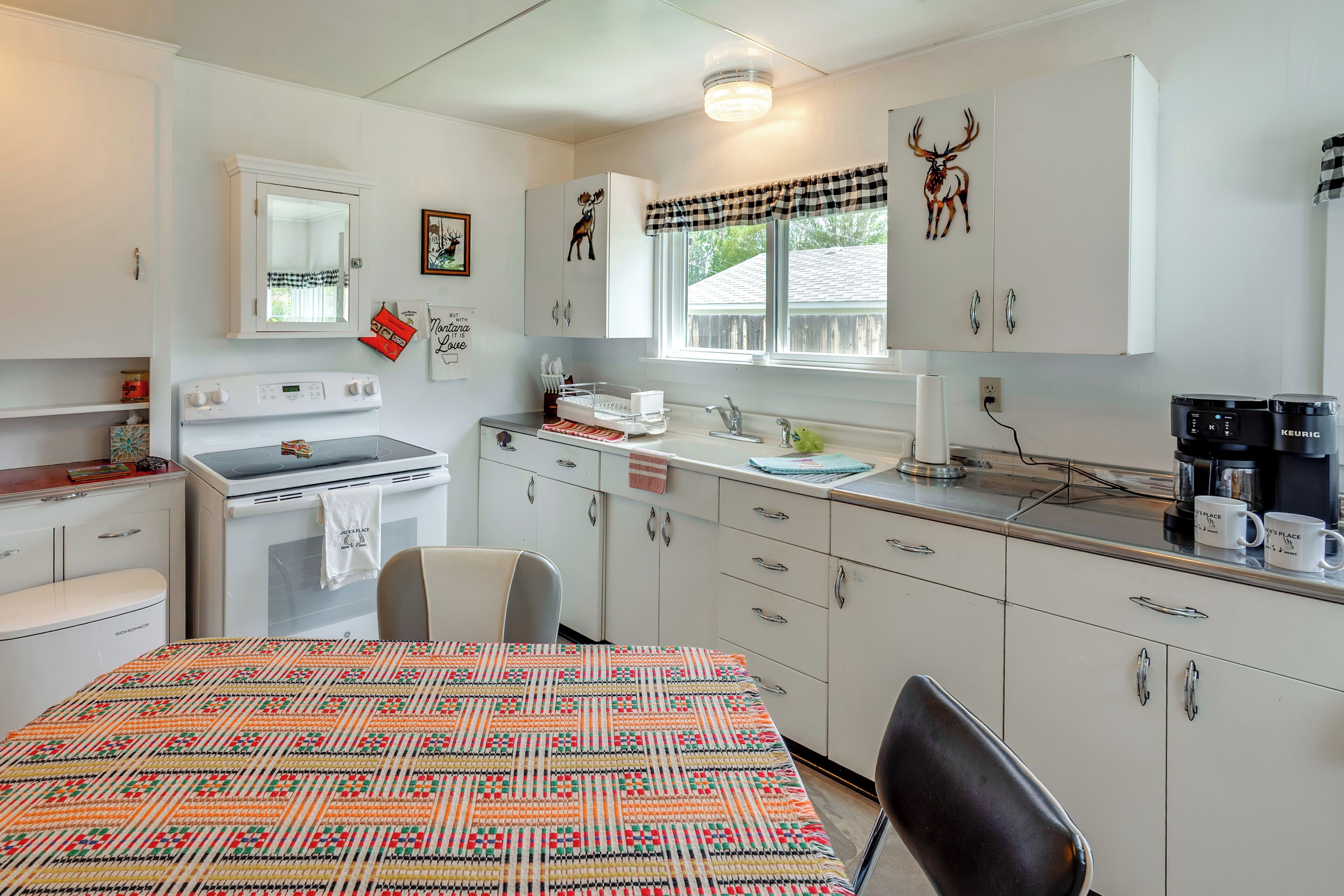A kitchen with white cabinets, a stove, a refrigerator, a window with a checkered valance, and a wall-mounted deer decoration.