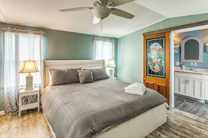 A bedroom with a bed, a ceiling fan, a nightstand with a lamp, a door with a decorative glass panel, and a bathroom with a sink and mirror.