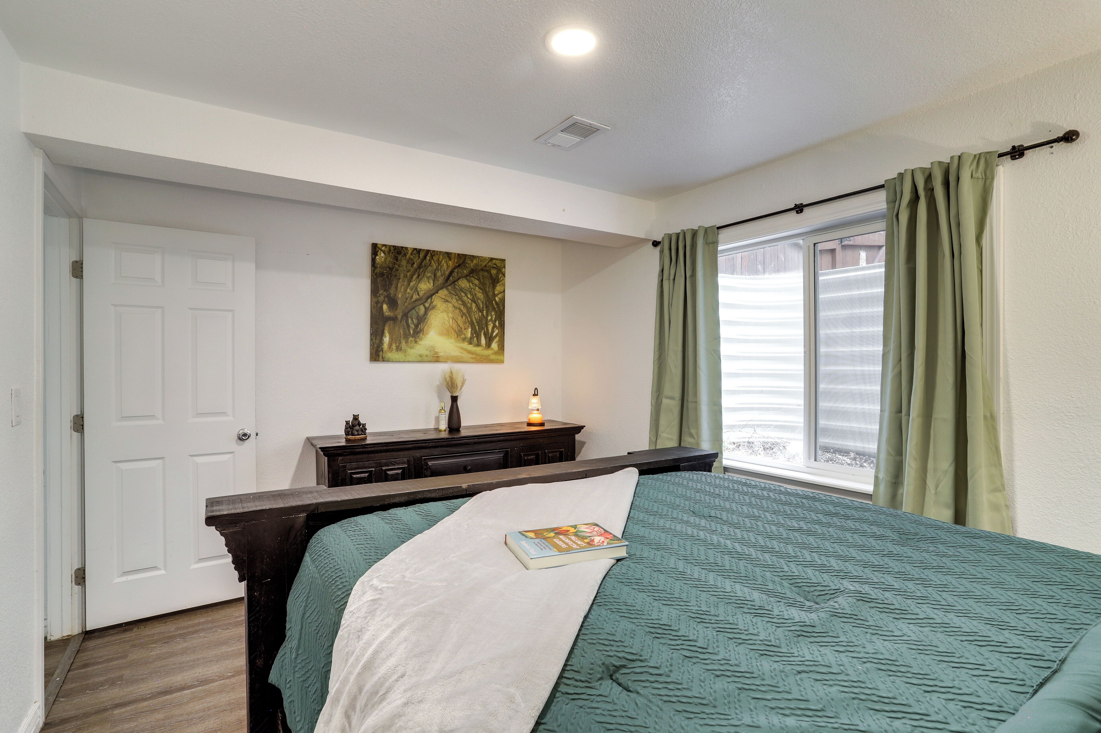 A bedroom with a bed, a nightstand, a painting, a window with curtains, and a door.