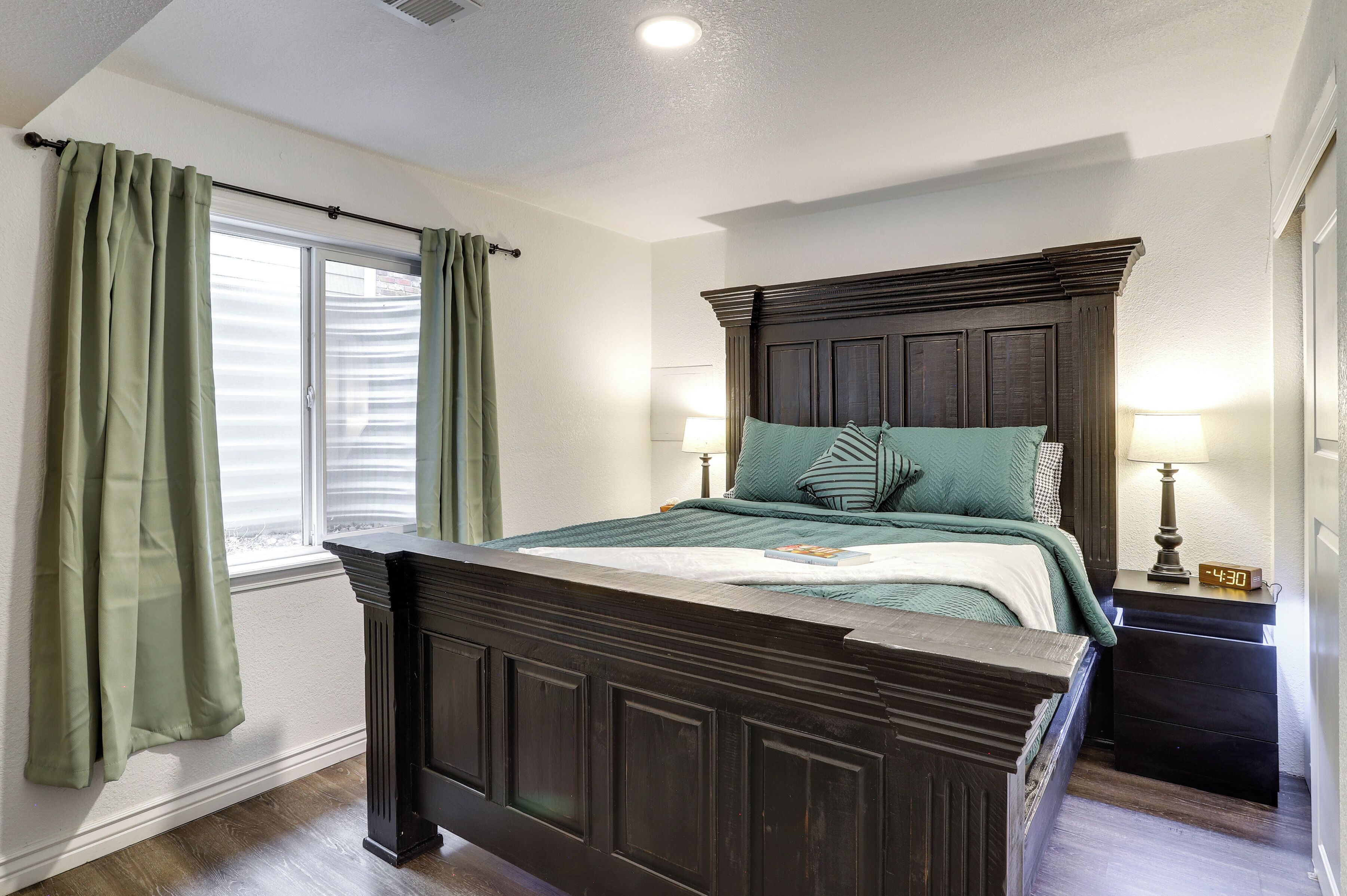 A bedroom with a dark wooden bed, a window with green curtains, a bedside lamp, and a clock.