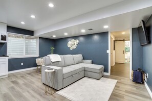 Uma sala de estar com um sofá seccional, uma pequena mesa lateral, um objeto decorativo na parede e uma televisão fixada na parede.