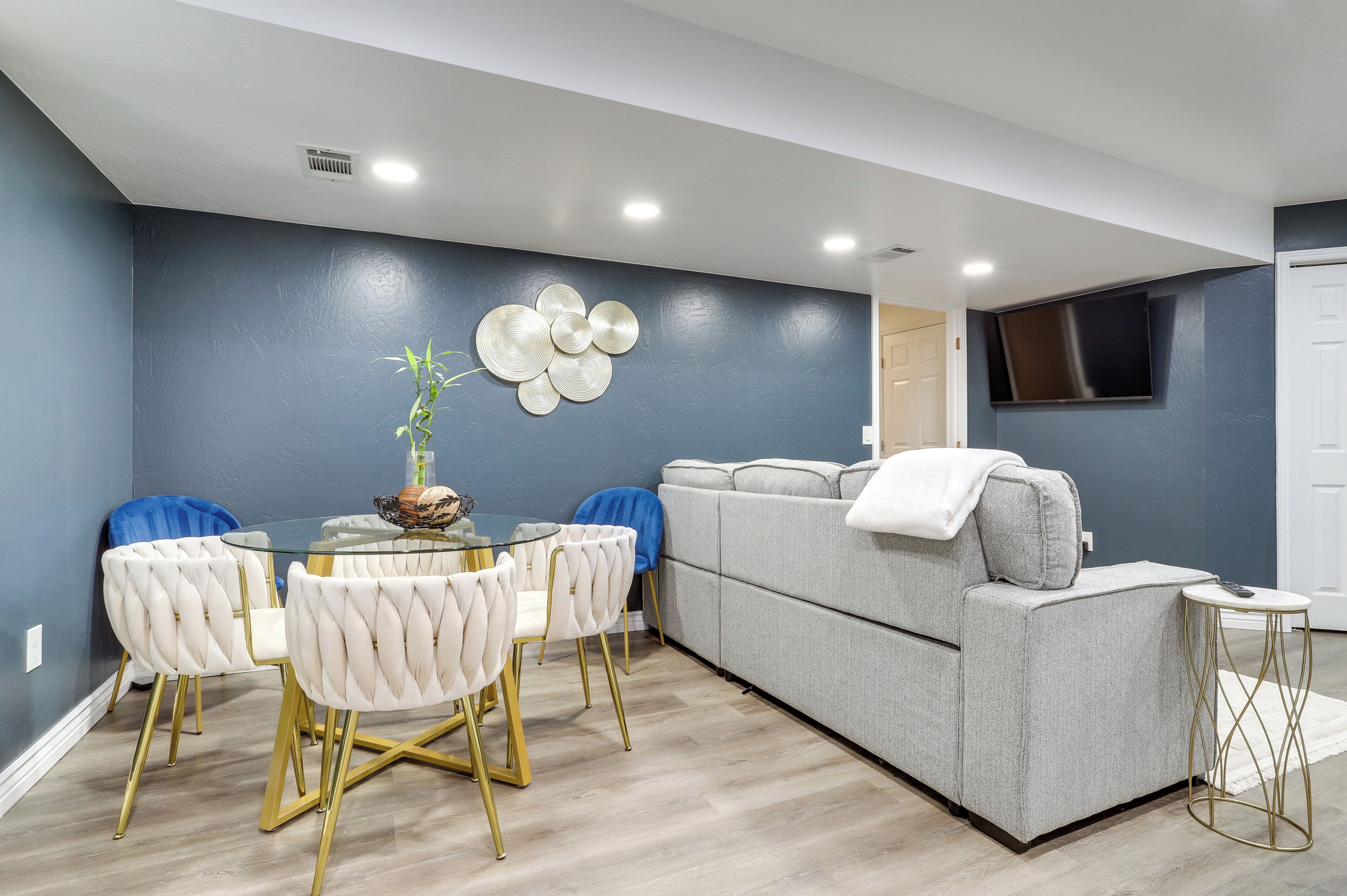 A modern living room with a grey sofa, a glass dining table with white upholstered chairs, and a decorative wall piece.