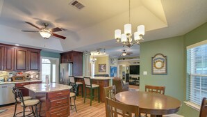 A spacious kitchen with wooden cabinets, a central island with bar stools, a dining table with chairs, and a ceiling fan. - Pool & Covered Patio! Galveston County Getaway (Dickinson)