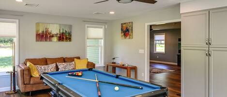 A pool table with pool cues and balls, a brown leather sofa with cushions, a ceiling fan, and artwork on the walls.