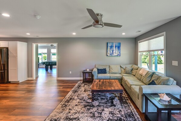 A spacious living room with a ceiling fan, a light-colored sofa, a coffee table, and a large window with blinds.